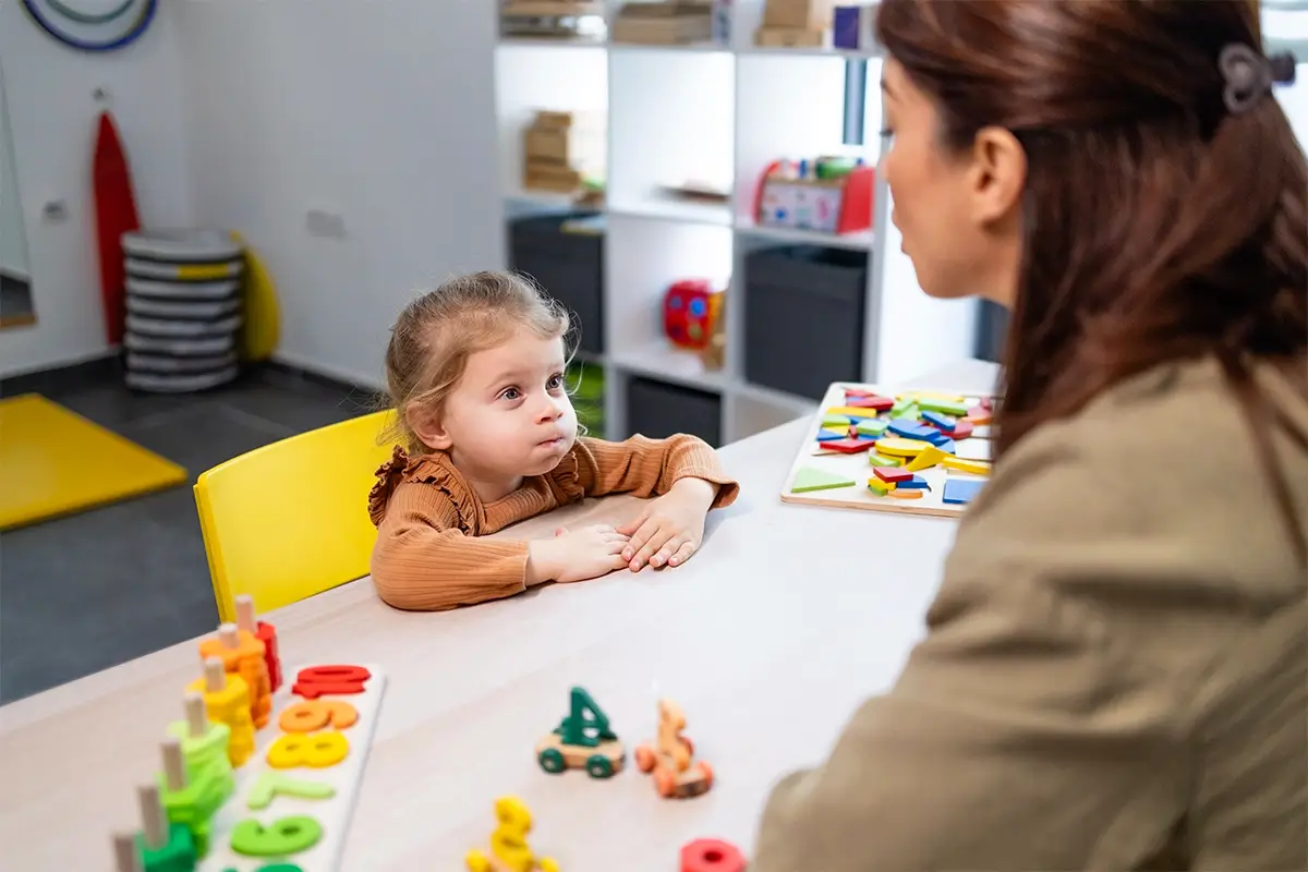 A young child sits at a table with colorful toys and looks attentively at an adult woman, illustrating how a child won't always listen to parents but may respond differently to other trusted adults.