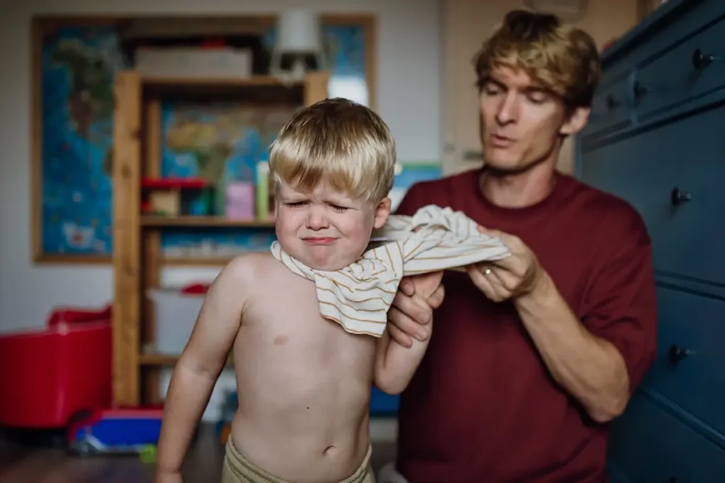 A father calmly tries to dress his resistant young son, who grimaces and pulls away — an everyday example of why emotional regulation is hard for kids and how co-regulation begins with a patient parent.