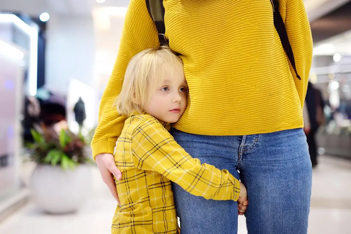 A young child clings tightly to a parent's leg in a busy public space, reflecting the anxiety and need for security that can underlie why kids won't listen to parents in overwhelming environments.