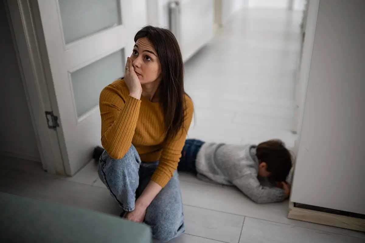 A worn-out mother sits on the floor with her hand to her face while her child lies face-down in the hallway mid-meltdown, reflecting the exhaustion parents feel when kids struggle with emotional regulation.