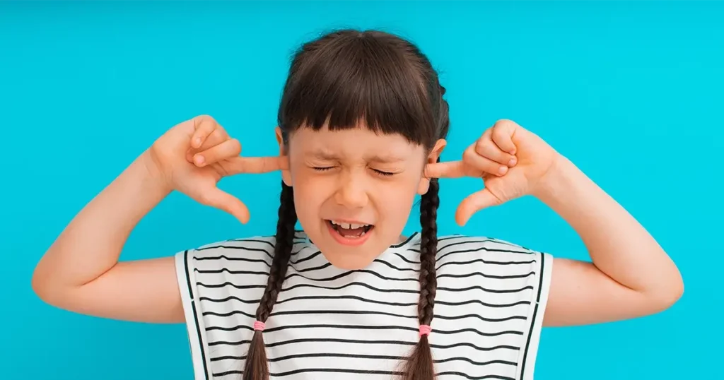 A young girl with braided pigtails and wearing a striped t-shirt puts her fingers in her ears and squeezes her eyes shut, illustrating why kids don't listen to parents.