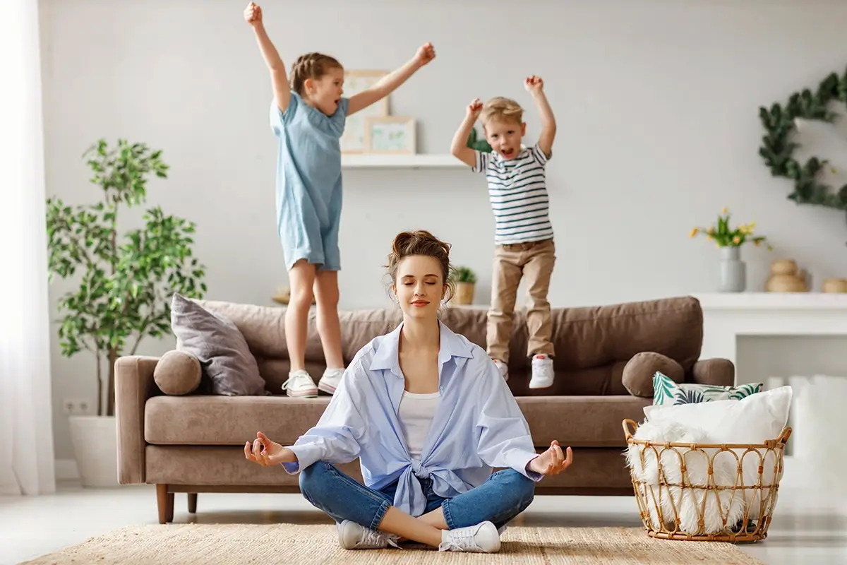 A mother sits in a calm meditation pose on the living room floor while two children jump and shout on the sofa behind her, illustrating the challenge of parent emotional regulation amid household chaos.