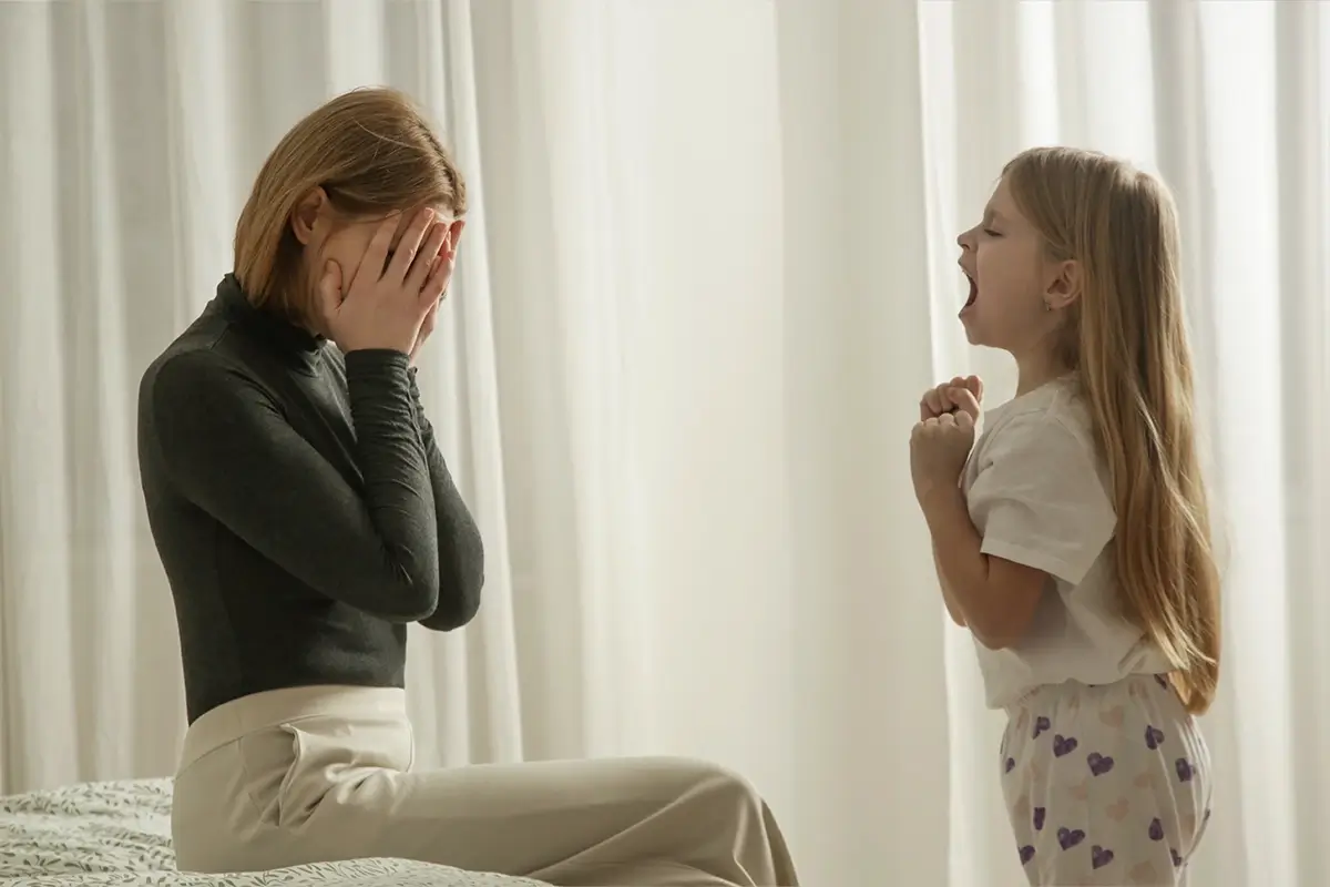 A stressed mother sits on a bed with her face buried in her hands while her young daughter shouts at her, capturing the moment when a parent's own emotional regulation is pushed to its limit.