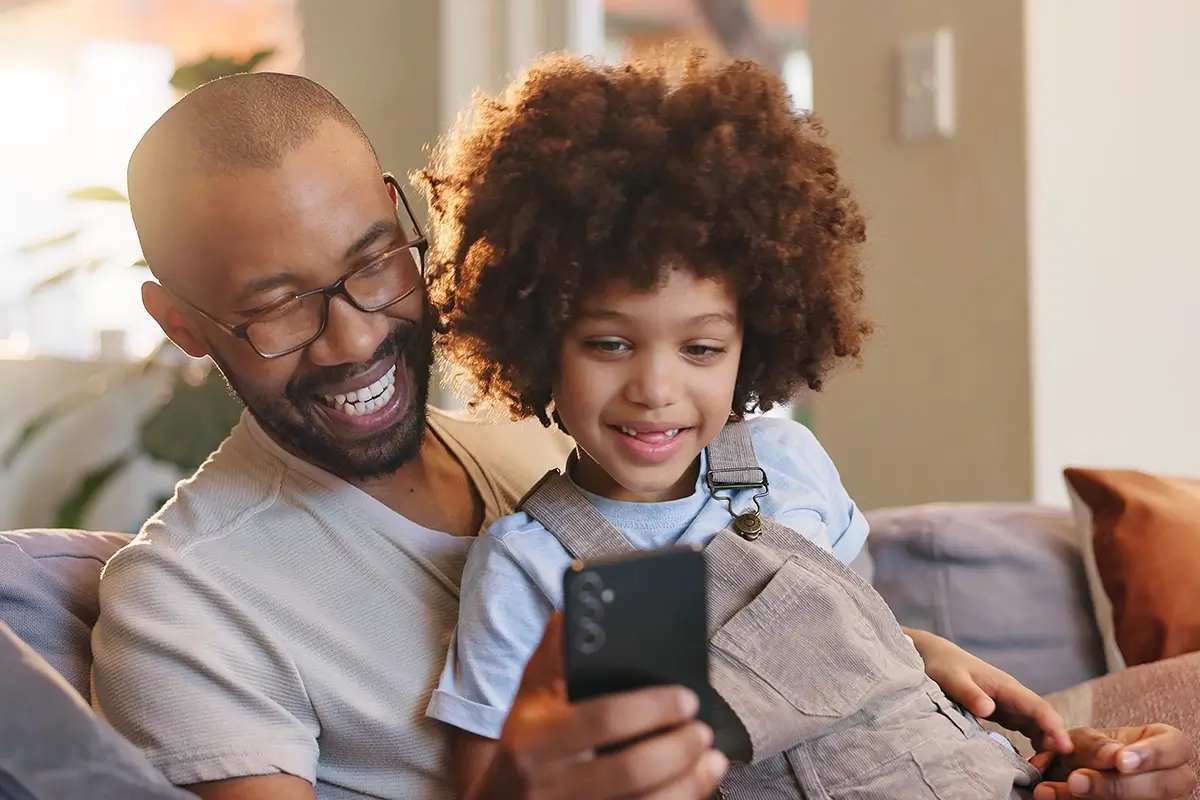A father and young child sit together on a sofa, both smiling as they look at a smartphone, showing how the right approach can turn a moment of resistance into a positive connection between parent and child.