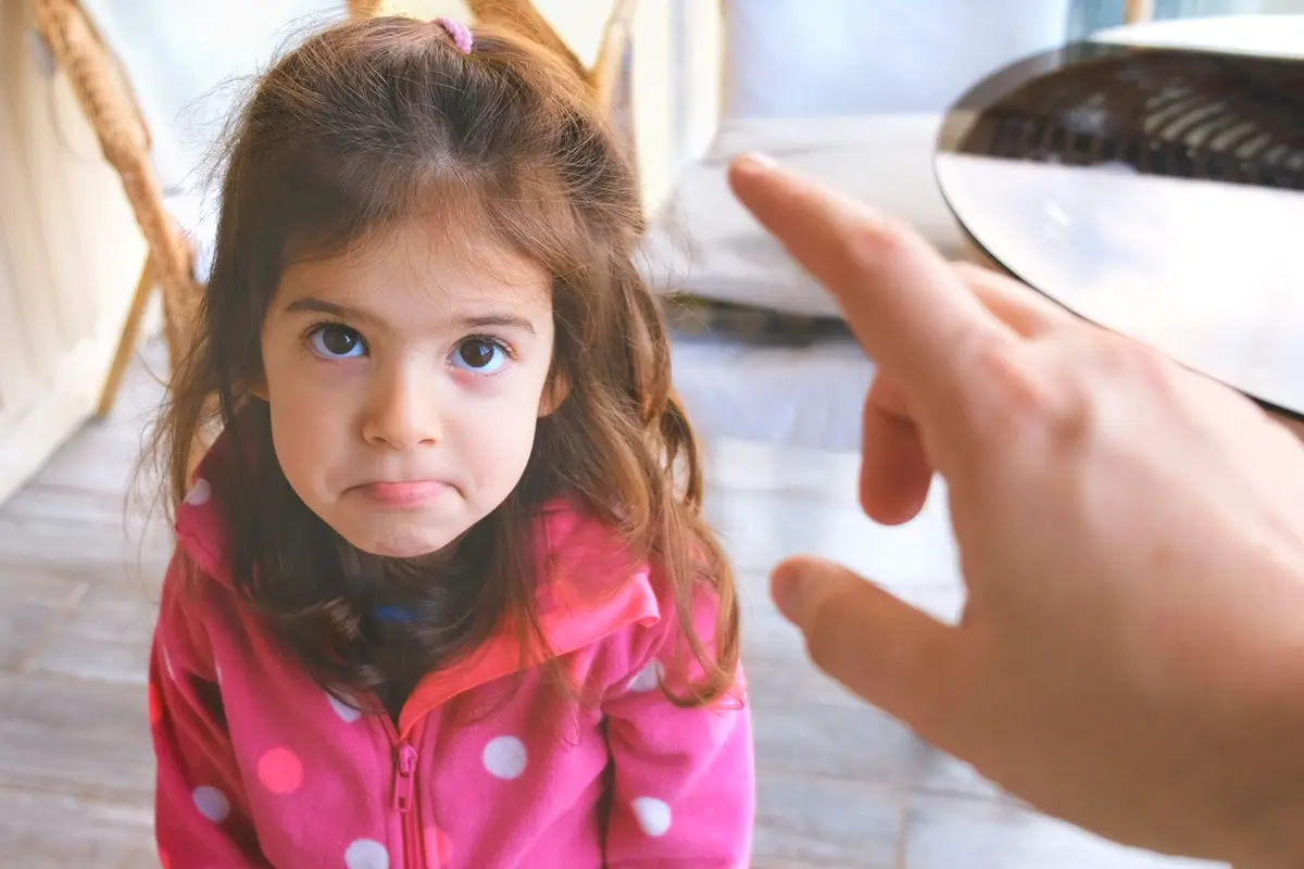 A young girl looks up with a defiant expression as an adult points a finger toward her, depicting the standoff many parents experience when their child won't listen to instructions.