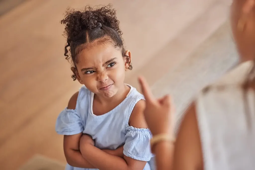 A young girl crosses her arms and scowls up at an adult who is pointing at her, illustrating the emotional dysregulation and defiance that can precede a meltdown or tantrum in young children.