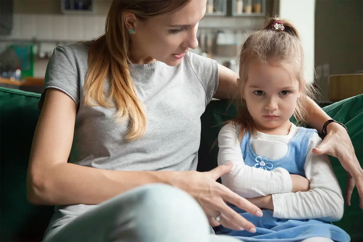 A mother speaks gently to her young daughter on a sofa, while the child sits with arms crossed and a stubborn expression — capturing the frustration parents feel when a child won't listen.